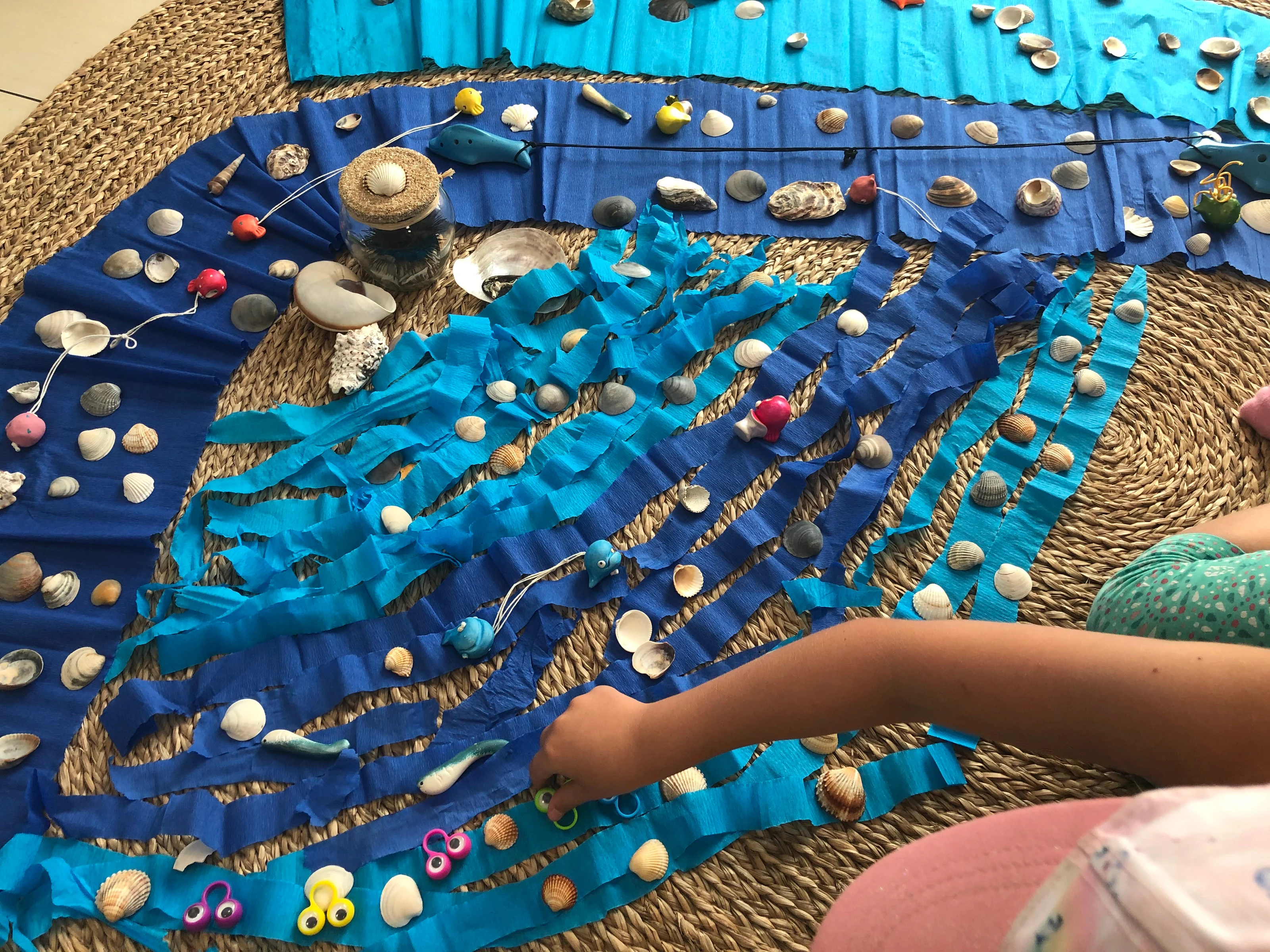 Auf einem runden Bastteppich ist aus blauen Krepppapierbahnen ein Meer gelegt, belegt mit Muscheln und kleinen Fischen; ein Kind greift von rechts hinein.