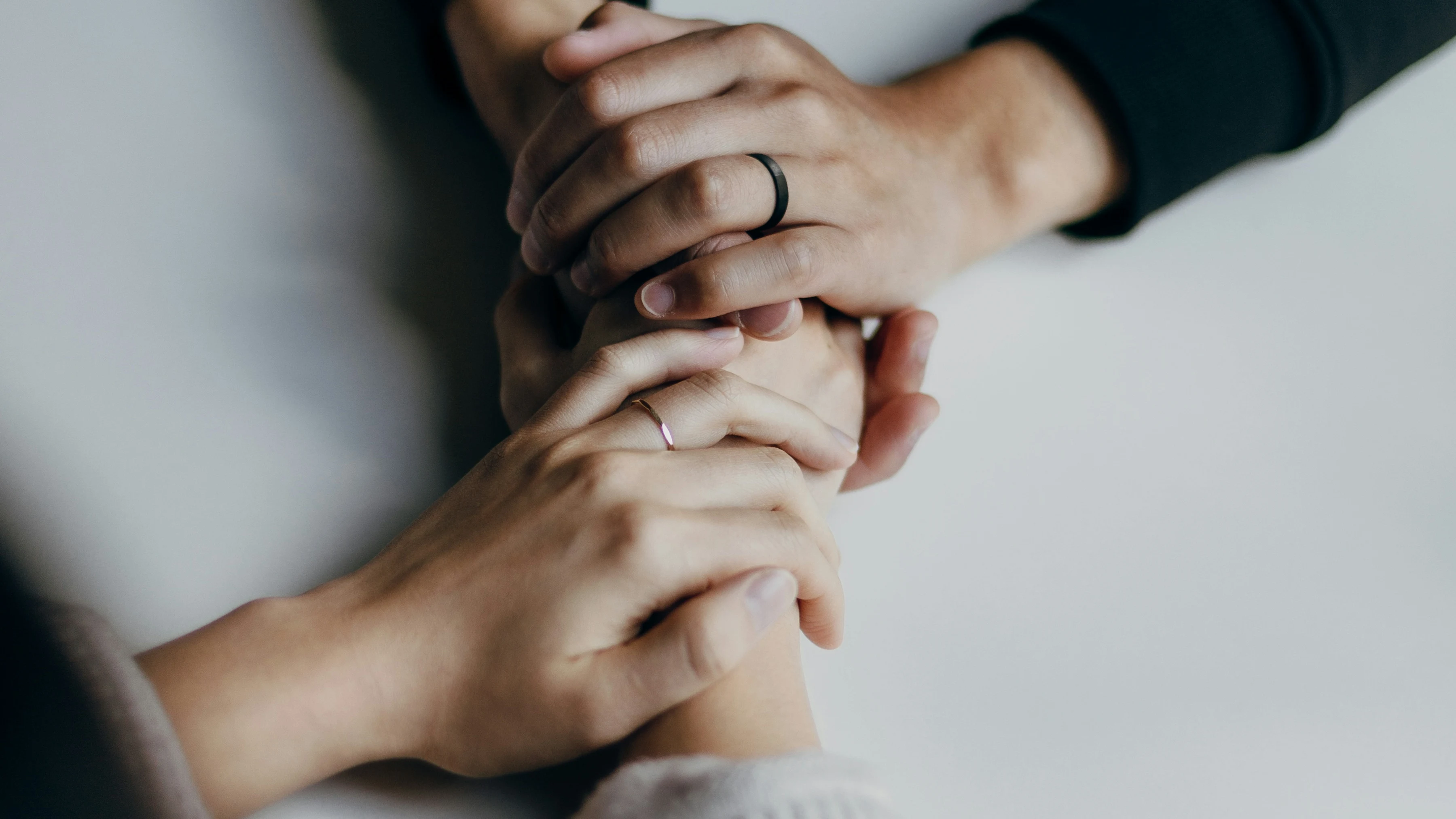 Über einer hellen Tischplatte halten zwei Menschen einander mit beiden Händen fest; beide tragen einen Ring.
