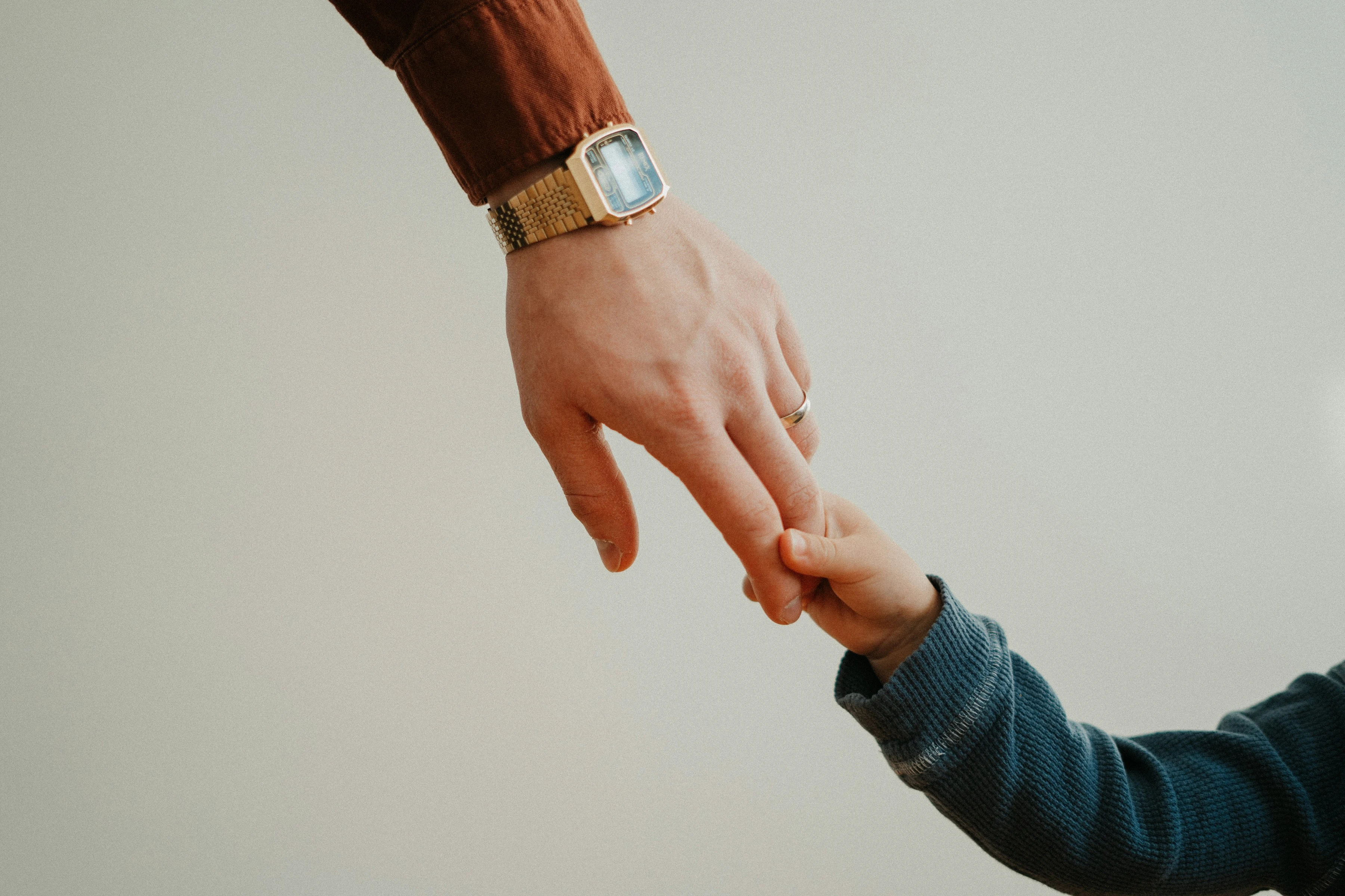 Eine Kinderhand umfasst den Zeigefinger einer erwachsenen Hand; der Erwachsene trägt einen terrakottafarbenen Ärmel und eine Armbanduhr, das Kind einen blauen Strickpullover.