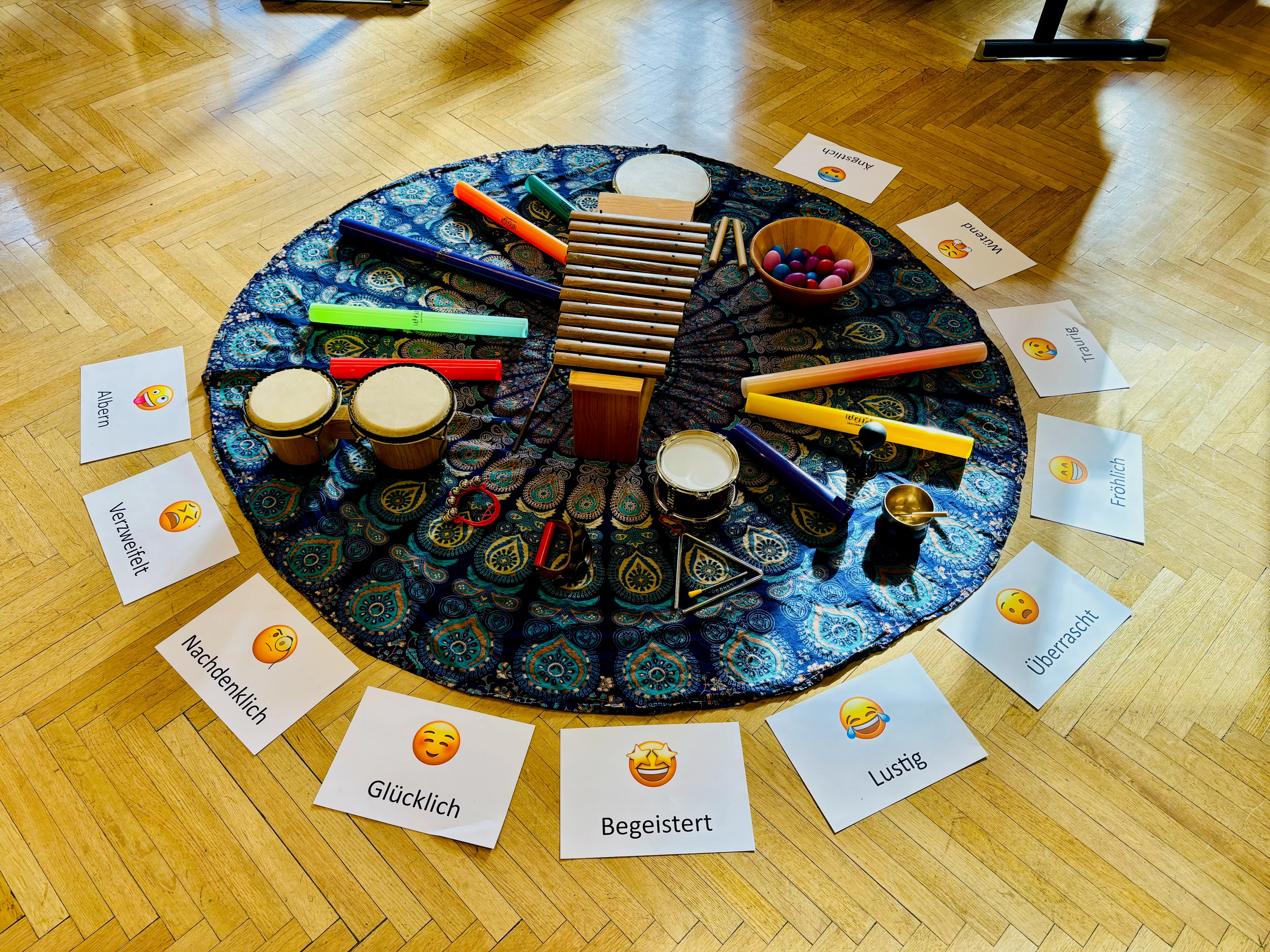 Auf einem runden blauen Mandalatuch liegen Instrumente – Xylophon, Bongos, Trommel, Triangel, Klangröhren und eine Klangschale. Ringsum auf dem Parkett liegen Karten mit Gefühlen: glücklich, begeistert, lustig, traurig, wütend, ängstlich.